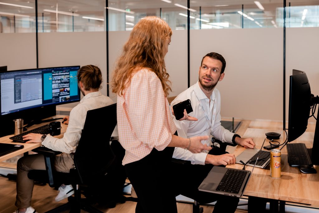 kvinne staar ved pulten til mann som sitter foran pc, de ser paa hverandre og snakker.